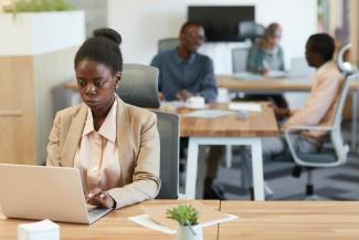 woman works on her laptop in a modern office, with colleagues in the background.